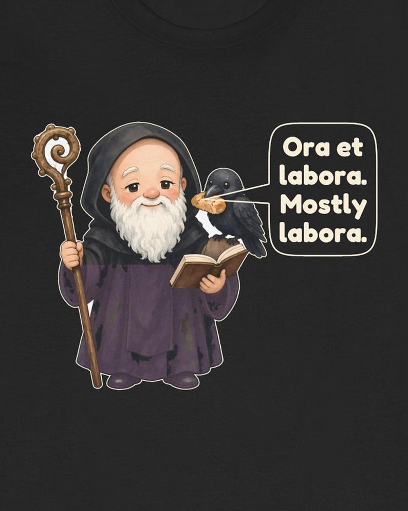 May include: A black t-shirt featuring a cartoon monk with a white beard and brown staff, holding an open book. A black raven sits on the book holding a piece of bread. A speech bubble contains the text: Ora et labora. Mostly labora.