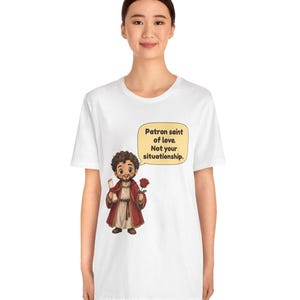 Puede incluir: Una camiseta blanca con una ilustraci&oacute;n de dibujos animados de una figura barbuda con t&uacute;nica roja que sostiene un pergamino y una rosa roja. Un bocadillo amarillo sobre la figura contiene el texto: Patron saint of love. Not your situationship.