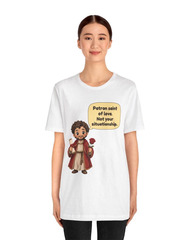 Puede incluir: Una camiseta blanca con una ilustraci&oacute;n de dibujos animados de una figura barbuda con t&uacute;nica roja que sostiene un pergamino y una rosa roja. Un bocadillo amarillo sobre la figura contiene el texto: Patron saint of love. Not your situationship.