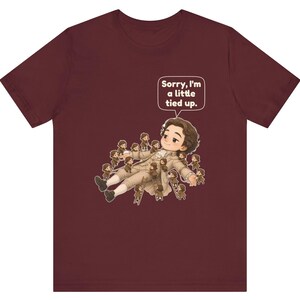 Op de afbeelding: Een bordeauxrood t-shirt met korte mouwen en een afbeelding van een groot personage dat ligt en omringd is door vele kleine figuurtjes op ladders. Een tekstballon boven het grote personage bevat de tekst, Sorry, I'm a little tied up.
