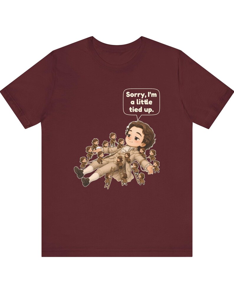Op de afbeelding: Een bordeauxrood t-shirt met korte mouwen en een afbeelding van een groot personage dat ligt en omringd is door vele kleine figuurtjes op ladders. Een tekstballon boven het grote personage bevat de tekst, Sorry, I'm a little tied up.