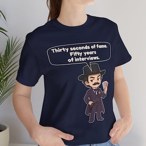 Pu&ograve; includere: Una t-shirt blu navy con un'illustrazione a cartone animato di un uomo con i baffi in abito scuro, cappello e distintivo da sceriffo, che tiene in mano delle carte da gioco. Un fumetto sopra il personaggio contiene il testo: Thirty seconds of fame. Fifty years of interviews.
