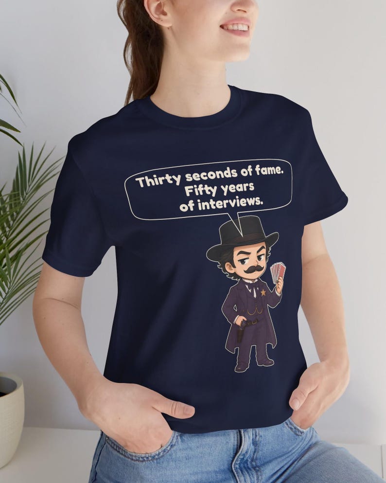 Pu&ograve; includere: Una t-shirt blu navy con un'illustrazione a cartone animato di un uomo con i baffi in abito scuro, cappello e distintivo da sceriffo, che tiene in mano delle carte da gioco. Un fumetto sopra il personaggio contiene il testo: Thirty seconds of fame. Fifty years of interviews.