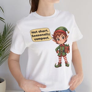 May include: A white short-sleeved t-shirt featuring a graphic of a cartoon elf holding a gift. A yellow speech bubble next to the elf contains the black text: Not short. Seasonally compact. The elf wears a green hat, green tunic, and red and white striped leggings.