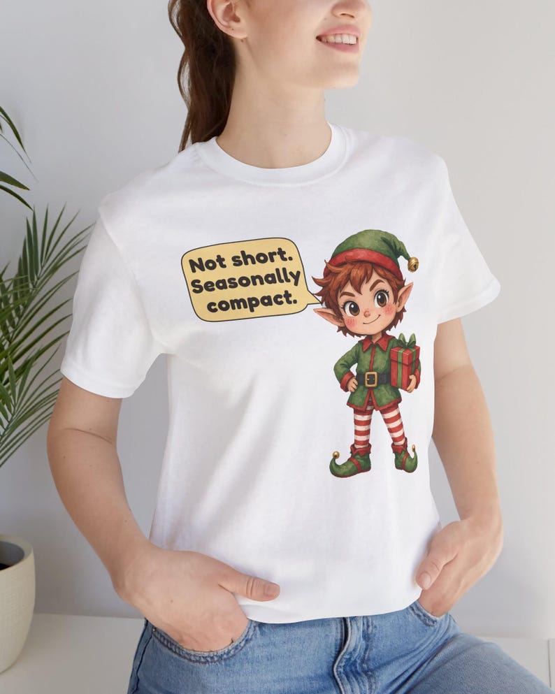 May include: A white short-sleeved t-shirt featuring a graphic of a cartoon elf holding a gift. A yellow speech bubble next to the elf contains the black text: Not short. Seasonally compact. The elf wears a green hat, green tunic, and red and white striped leggings.