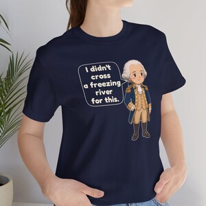 May include: A navy blue t-shirt featuring a cartoon illustration of George Washington in a colonial uniform. A speech bubble next to the figure contains the text: I didn't cross a freezing river for this. The shirt has short sleeves and a crew neckline.