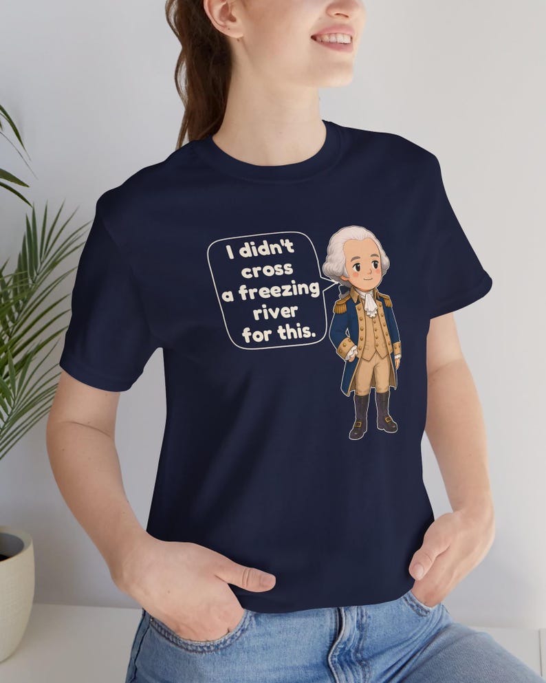 May include: A navy blue t-shirt featuring a cartoon illustration of George Washington in a colonial uniform. A speech bubble next to the figure contains the text: I didn't cross a freezing river for this. The shirt has short sleeves and a crew neckline.