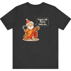 May include: A dark heather gray t-shirt featuring a cartoon illustration of a bearded figure in a red and yellow robe wearing a party hat, holding a sickle and wheat, with a speech bubble that reads, "Togas off. Party hats on."