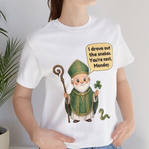 Puede incluir: Una camiseta blanca con una ilustraci&oacute;n de dibujos animados de una figura barbuda con vestimentas religiosas verdes, sosteniendo un bast&oacute;n de madera y un tr&eacute;bol. Una peque&ntilde;a serpiente verde est&aacute; al lado. Un globo de texto contiene: I drove out the snakes. You're next, Monday.