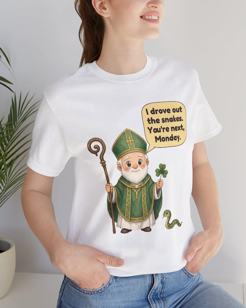 Puede incluir: Una camiseta blanca con una ilustraci&oacute;n de dibujos animados de una figura barbuda con vestimentas religiosas verdes, sosteniendo un bast&oacute;n de madera y un tr&eacute;bol. Una peque&ntilde;a serpiente verde est&aacute; al lado. Un globo de texto contiene: I drove out the snakes. You're next, Monday.