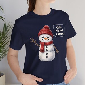 Pode incluir: Uma t-shirt azul-marinho com o gr&aacute;fico de um boneco de neve a usar um gorro e um cachecol de malha vermelhos. O boneco tem um nariz de cenoura, bra&ccedil;os de pau e bot&otilde;es pretos. Um bal&atilde;o de fala cont&eacute;m: Chill. It's just a phase.