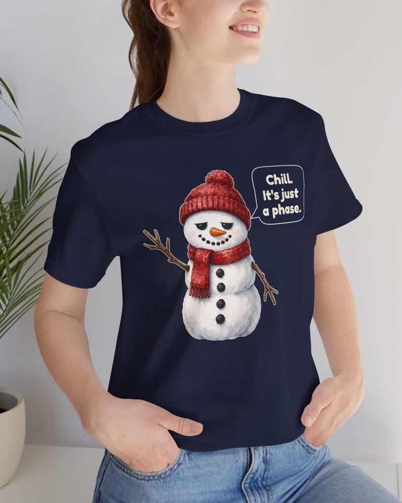 Pode incluir: Uma t-shirt azul-marinho com o gr&aacute;fico de um boneco de neve a usar um gorro e um cachecol de malha vermelhos. O boneco tem um nariz de cenoura, bra&ccedil;os de pau e bot&otilde;es pretos. Um bal&atilde;o de fala cont&eacute;m: Chill. It's just a phase.
