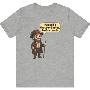 May include: A heather gray t-shirt featuring a cartoon illustration of a bearded traveler wearing a brown hat and coat, holding a walking stick. A speech bubble above the character contains the text: I walked a thousand miles. Pack a lunch.