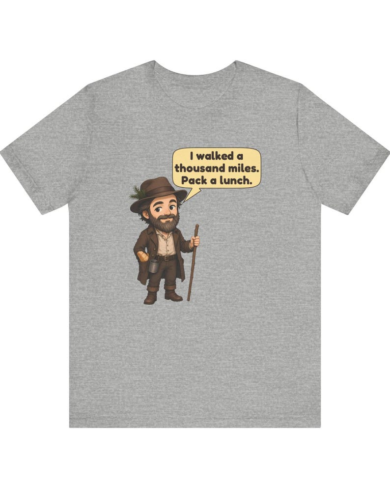 May include: A heather gray t-shirt featuring a cartoon illustration of a bearded traveler wearing a brown hat and coat, holding a walking stick. A speech bubble above the character contains the text: I walked a thousand miles. Pack a lunch.