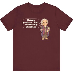 Pu&ograve; includere: Una t-shirt a maniche corte color bordeaux con un'illustrazione a cartone animato di un uomo con capelli bianchi, occhiali e cappotto viola che tiene in mano una piuma e una pergamena. Un fumetto accanto al personaggio contiene il testo: Hebrew professor. Poet. Accidentally Christmas.