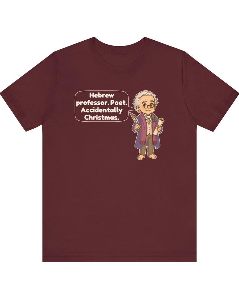 Pu&ograve; includere: Una t-shirt a maniche corte color bordeaux con un'illustrazione a cartone animato di un uomo con capelli bianchi, occhiali e cappotto viola che tiene in mano una piuma e una pergamena. Un fumetto accanto al personaggio contiene il testo: Hebrew professor. Poet. Accidentally Christmas.