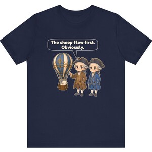 May include: A navy blue t-shirt featuring a graphic of two figures in 18th-century attire standing next to a hot air balloon with a sheep in the basket. A speech bubble above them contains the text: The sheep flew first. Obviously.