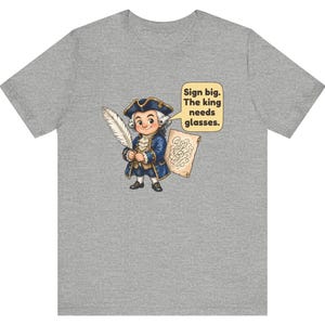May include: A heather gray t-shirt featuring a cartoon illustration of a person in 18th-century attire holding a quill and a scroll. A speech bubble contains the text: Sign big. The king needs glasses.