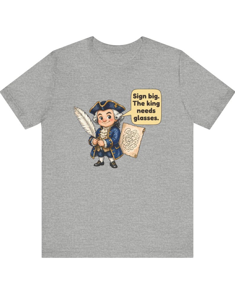 May include: A heather gray t-shirt featuring a cartoon illustration of a person in 18th-century attire holding a quill and a scroll. A speech bubble contains the text: Sign big. The king needs glasses.