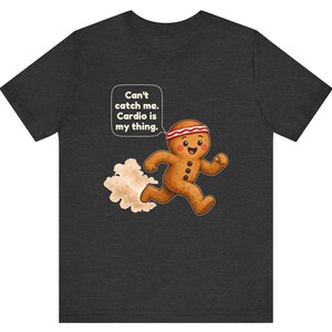 May include: A dark charcoal grey t-shirt featuring a graphic of a running gingerbread person wearing a red and white headband. A speech bubble above the character contains the text: Can't catch me. Cardio is my thing. A dust cloud follows.
