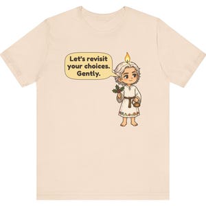 May include: A light beige t-shirt featuring a graphic of a cartoon character with a candle flame on their head, holding a sprig of holly and a small kettle. A speech bubble contains the text: Let's revisit your choices. Gently.