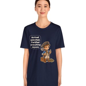 May include: A navy blue t-shirt featuring a cartoon illustration of a historical figure in a blue coat and tricorn hat holding a shovel and compass, with a wooden chest and text that reads: Arrived uninvited. Fortified everything anyway.
