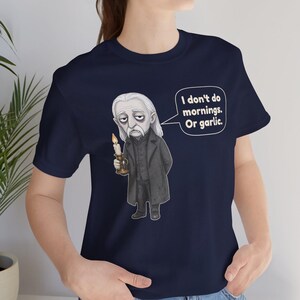 May include: A navy blue short-sleeve t-shirt featuring a graphic illustration of a pale, long-haired character in a dark coat holding a lit candle. A speech bubble next to the character contains the text: I don't do mornings. Or garlic.