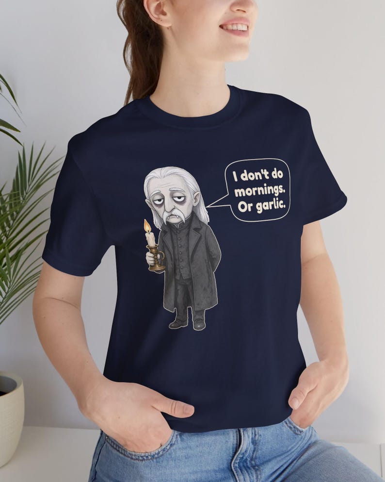 May include: A navy blue short-sleeve t-shirt featuring a graphic illustration of a pale, long-haired character in a dark coat holding a lit candle. A speech bubble next to the character contains the text: I don't do mornings. Or garlic.