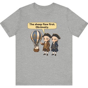 May include: A heather gray t-shirt featuring a graphic of two historical figures in tricorn hats standing next to a hot air balloon carrying a sheep. A speech bubble above them contains the text, "The sheep flew first. Obviously."