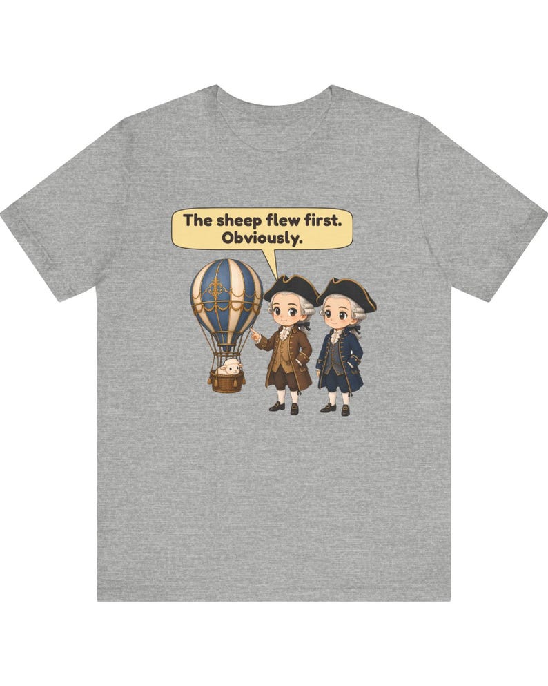 May include: A heather gray t-shirt featuring a graphic of two historical figures in tricorn hats standing next to a hot air balloon carrying a sheep. A speech bubble above them contains the text, "The sheep flew first. Obviously."