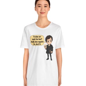 以下が含まれることがあります： 白いTシャツに、黒い服を着て頭蓋骨を持っているキャラクターの漫画風イラストが描かれています。黄色い吹き出しには「To be or not to be? Ask me again in Act 5.」というテキストが含まれています。デザインはTシャツの前面中央に配置されています。