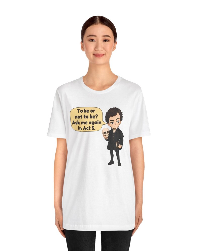 以下が含まれることがあります： 白いTシャツに、黒い服を着て頭蓋骨を持っているキャラクターの漫画風イラストが描かれています。黄色い吹き出しには「To be or not to be? Ask me again in Act 5.」というテキストが含まれています。デザインはTシャツの前面中央に配置されています。