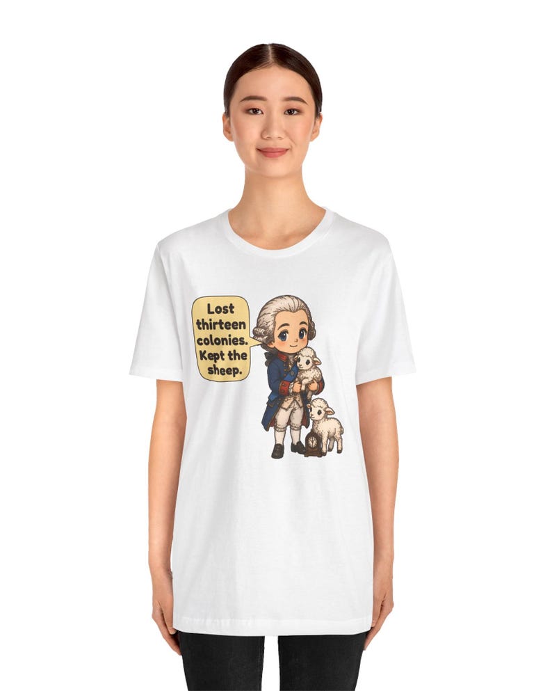 Peut inclure: Un t-shirt blanc avec une illustration de style dessin anim&eacute; repr&eacute;sentant un personnage historique en manteau bleu tenant un petit mouton blanc, avec un autre mouton &agrave; c&ocirc;t&eacute;. Une bulle contient le texte : Lost thirteen colonies. Kept the sheep.