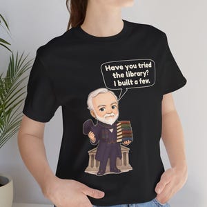 May include: A black t-shirt featuring a cartoon illustration of a bearded man in a suit holding a stack of books and a top hat. A speech bubble above him contains the text: Have you tried the library? I built a few. The man stands before a building.