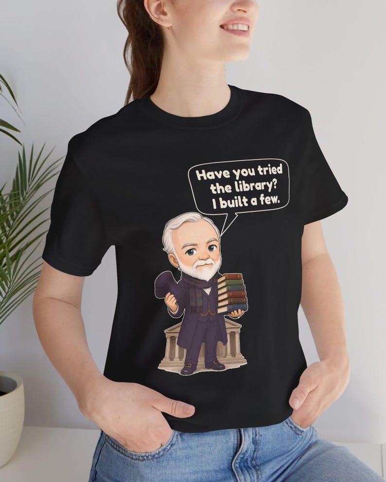 May include: A black t-shirt featuring a cartoon illustration of a bearded man in a suit holding a stack of books and a top hat. A speech bubble above him contains the text: Have you tried the library? I built a few. The man stands before a building.