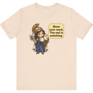 Puede incluir: Una camiseta de color beige claro con un gr&aacute;fico de un personaje con casco dorado y t&uacute;nica azul, sosteniendo una rama de olivo y un portapapeles. Un peque&ntilde;o b&uacute;ho se posa sobre el hombro junto a un bocadillo que dice: Show your work. The owl is watching.