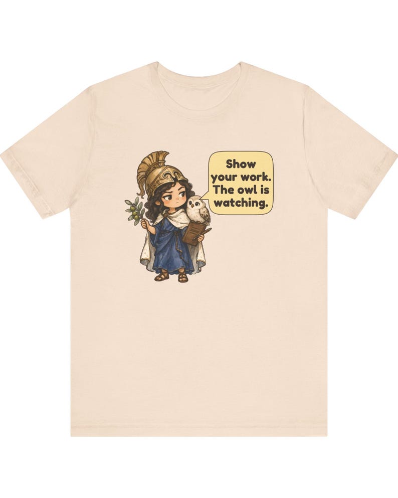 Puede incluir: Una camiseta de color beige claro con un gr&aacute;fico de un personaje con casco dorado y t&uacute;nica azul, sosteniendo una rama de olivo y un portapapeles. Un peque&ntilde;o b&uacute;ho se posa sobre el hombro junto a un bocadillo que dice: Show your work. The owl is watching.