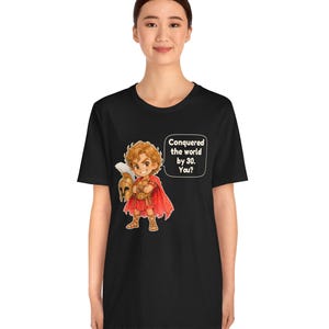 May include: A black t-shirt featuring a cartoon illustration of a historical figure in red armor and a cape, holding a helmet. A speech bubble next to the character contains the text: Conquered the world by 30. You?