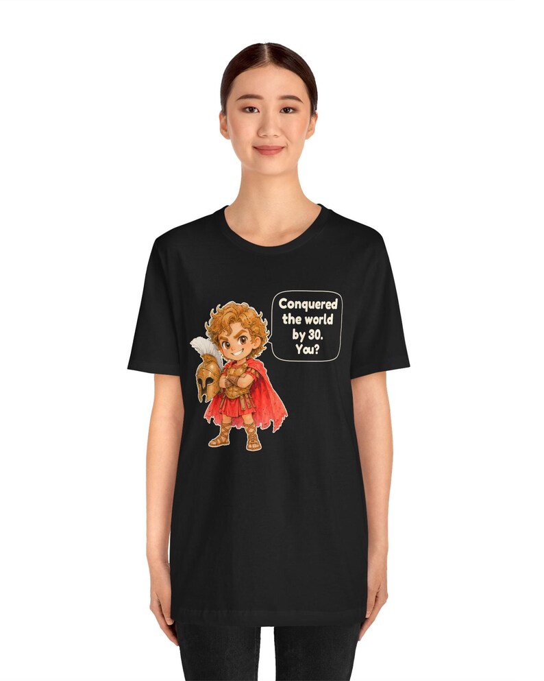 May include: A black t-shirt featuring a cartoon illustration of a historical figure in red armor and a cape, holding a helmet. A speech bubble next to the character contains the text: Conquered the world by 30. You?
