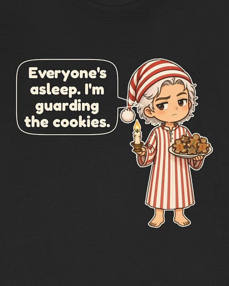 Op de afbeelding: Een zwart t-shirt met een stripfiguur in een rood-wit gestreepte pyjama en een bijpassende slaapmuts. Het figuur houdt een brandende kaars en een bord met peperkoekkoekjes vast. Een tekstballon bevat de tekst: Everyone's asleep. I'm guarding the cookies.