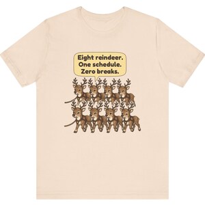 K&ouml;nnte beinhalten: Ein hellbeiges T-Shirt mit einer Grafik von acht braunen Rentieren in zwei Reihen, die durch Geschirre verbunden sind. &Uuml;ber den Rentieren befindet sich eine gelbe Sprechblase mit dem schwarzen Text: Eight reindeer. One schedule. Zero breaks.