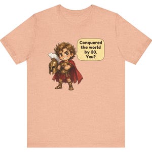 May include: A peach-colored t-shirt featuring a cartoon illustration of a warrior in armor holding a helmet, with a speech bubble containing the text: Conquered the world by 30. You? The warrior wears a red cape and brown sandals.