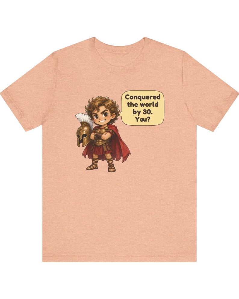 May include: A peach-colored t-shirt featuring a cartoon illustration of a warrior in armor holding a helmet, with a speech bubble containing the text: Conquered the world by 30. You? The warrior wears a red cape and brown sandals.