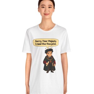 May include: A white t-shirt featuring a graphic of a cartoon figure wearing a black robe, a gold chain, and a halo. The figure holds a scroll and a book. A speech bubble above the figure contains the text: Sorry, Your Majesty. I read the fine print.