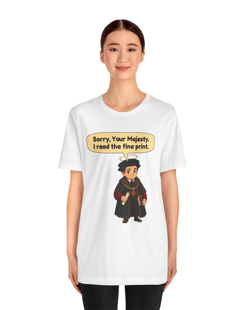 May include: A white t-shirt featuring a graphic of a cartoon figure wearing a black robe, a gold chain, and a halo. The figure holds a scroll and a book. A speech bubble above the figure contains the text: Sorry, Your Majesty. I read the fine print.