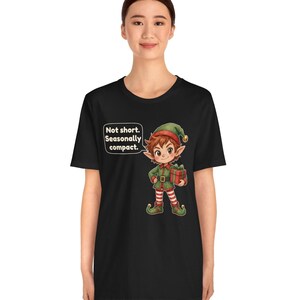 May include: A black t-shirt featuring a cartoon elf illustration holding a red gift box. The elf wears a green and red outfit with striped leggings. A speech bubble next to the elf contains the text: Not short. Seasonally compact.