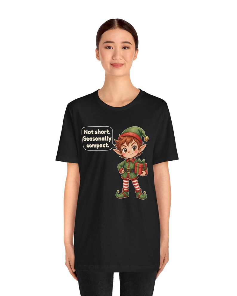 May include: A black t-shirt featuring a cartoon elf illustration holding a red gift box. The elf wears a green and red outfit with striped leggings. A speech bubble next to the elf contains the text: Not short. Seasonally compact.