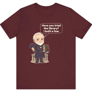 May include: A maroon short-sleeve t-shirt featuring a cartoon illustration of a bearded man in a suit holding a stack of books and a hat. A speech bubble above him contains the text: Have you tried the library? I built a few.