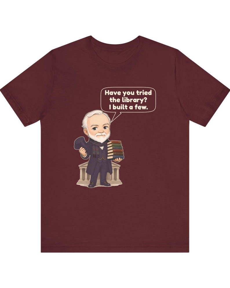 May include: A maroon short-sleeve t-shirt featuring a cartoon illustration of a bearded man in a suit holding a stack of books and a hat. A speech bubble above him contains the text: Have you tried the library? I built a few.