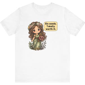 May include: A white t-shirt featuring a cartoon illustration of a girl with long brown hair wearing a flower crown and a green dress. She holds a pomegranate, and a speech bubble next to her contains the text: Six seeds. Totally worth it.